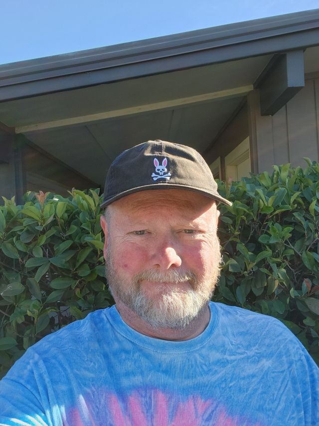 A man with a gray beard is standing outdoors, wearing a black cap with a bunny logo and a colorful tie-dye shirt, with green bushes and a house in the background.