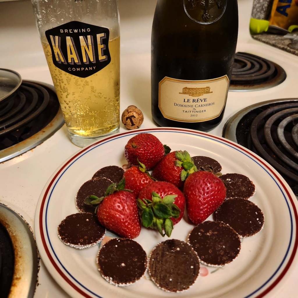 A plate with fresh strawberries and chocolate disks, accompanied by a glass of beer and a bottle of sparkling wine on a kitchen countertop.