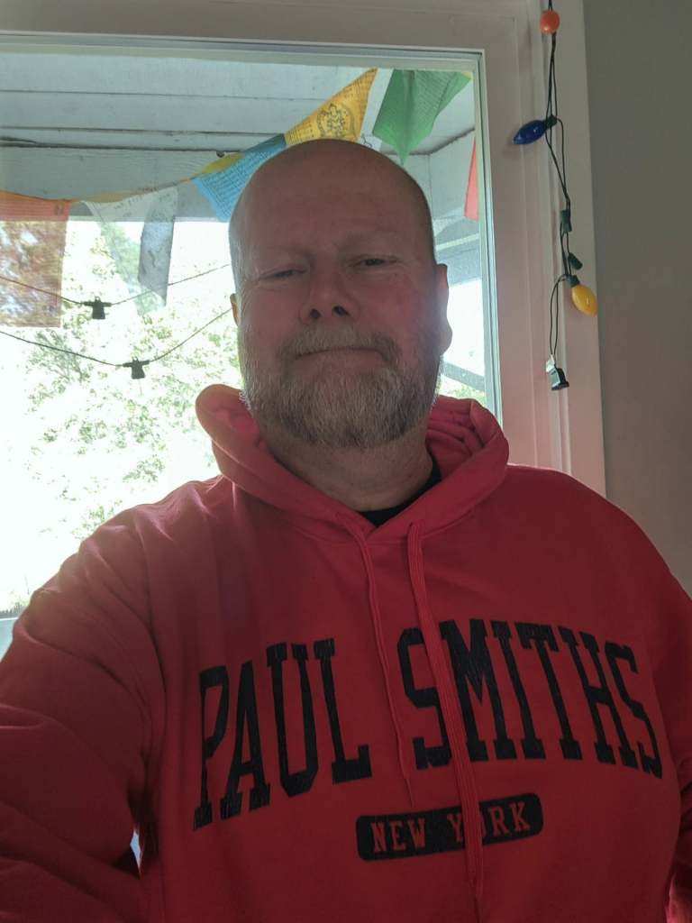 A person wearing a red hoodie with 'PAUL SMITHS NEW YORK' printed on it, standing in front of a window with decorative lights and colorful flags in the background.