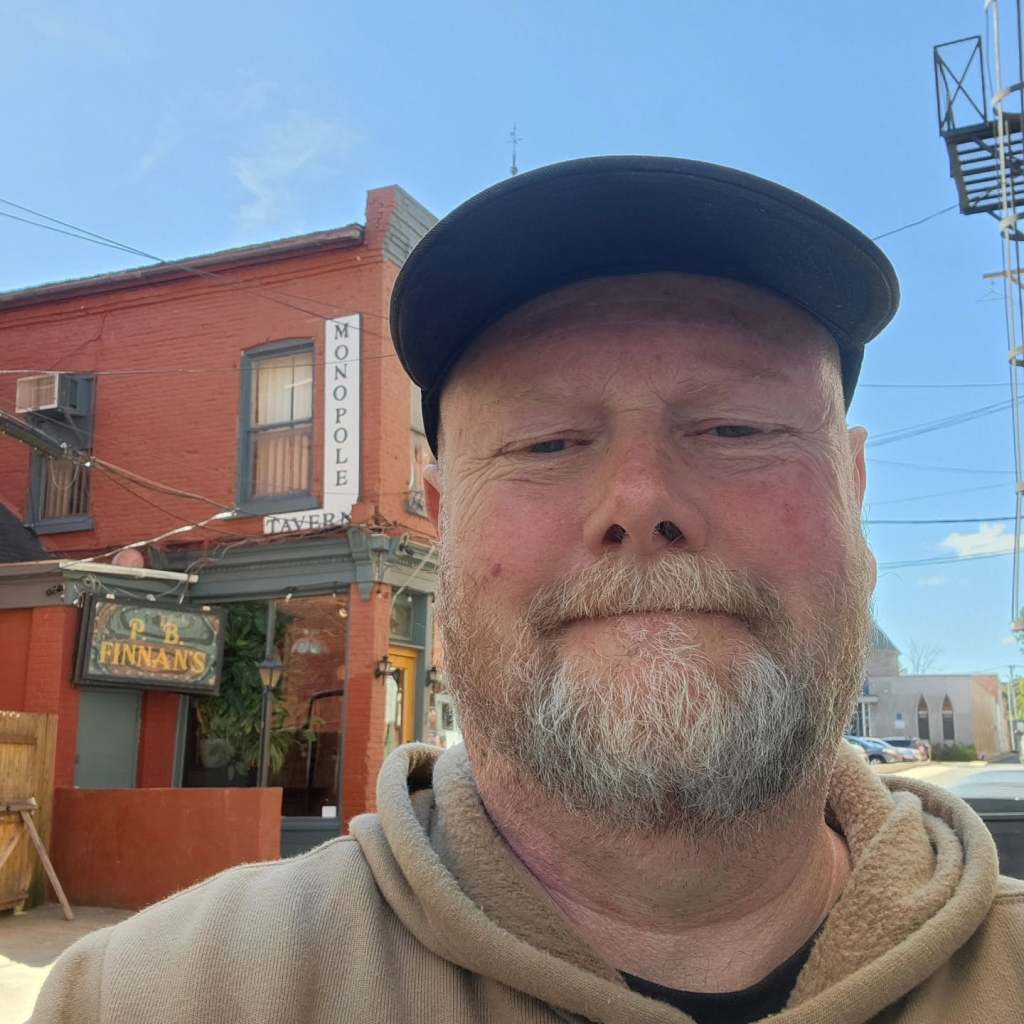 Selfie of a person with a beard wearing a cap, in front of a brick building with signs for 'Monopole Tavern' and 'P.B. Finnan's'.