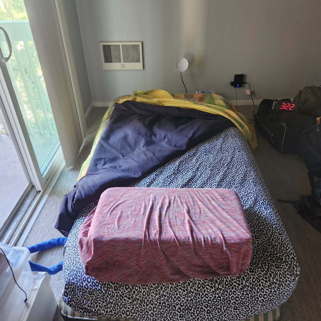 A neatly made bed with a patterned blanket, featuring a red pillow and a bedside lamp. There's a digital clock showing the time, alongside a backpack and storage container nearby. A large window allows natural light into the room.