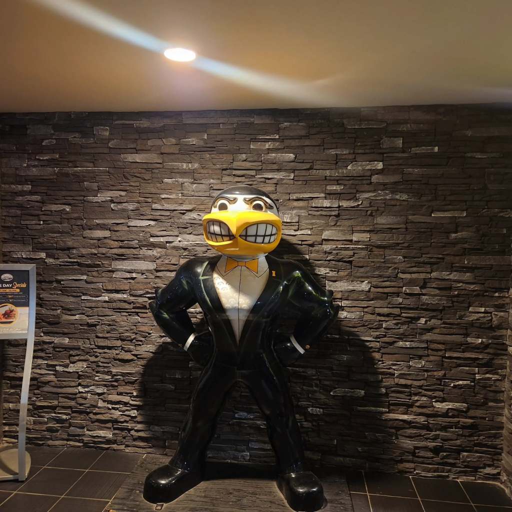 A large, cartoonish statue of a duck wearing a tuxedo, standing confidently in front of a stone wall.