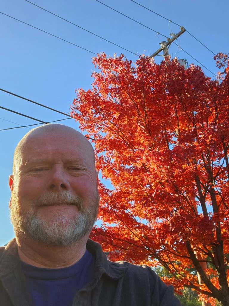 A man with a beard smiles for a selfie in front of a vibrant red autumn tree under a clear blue sky.