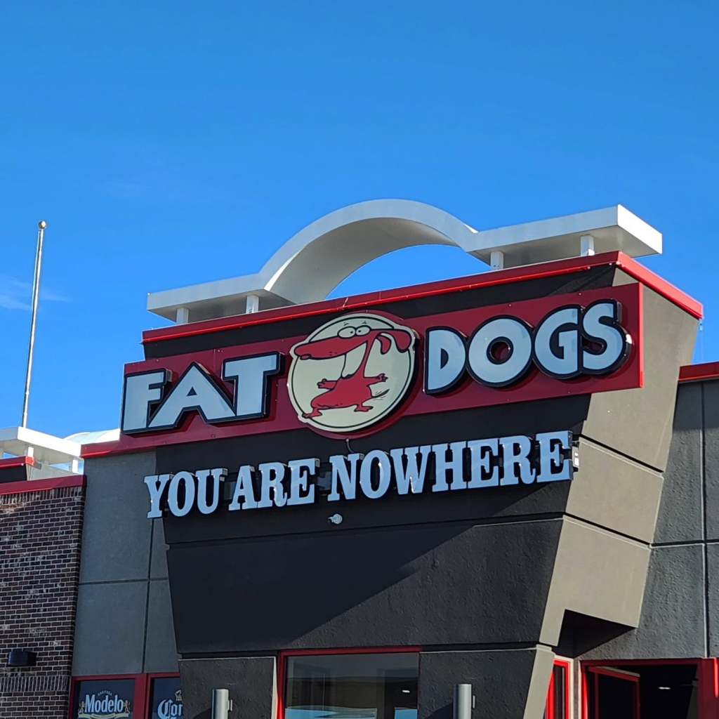 Sign of 'Fat Dogs' restaurant with the tagline 'YOU ARE NOWHERE' against a clear blue sky.