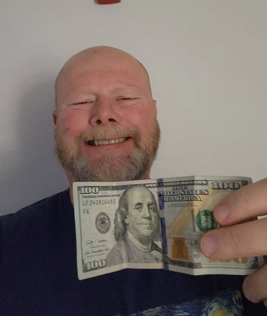 A smiling man with a beard holds a $100 bill in front of him, showcasing the front side of the currency.