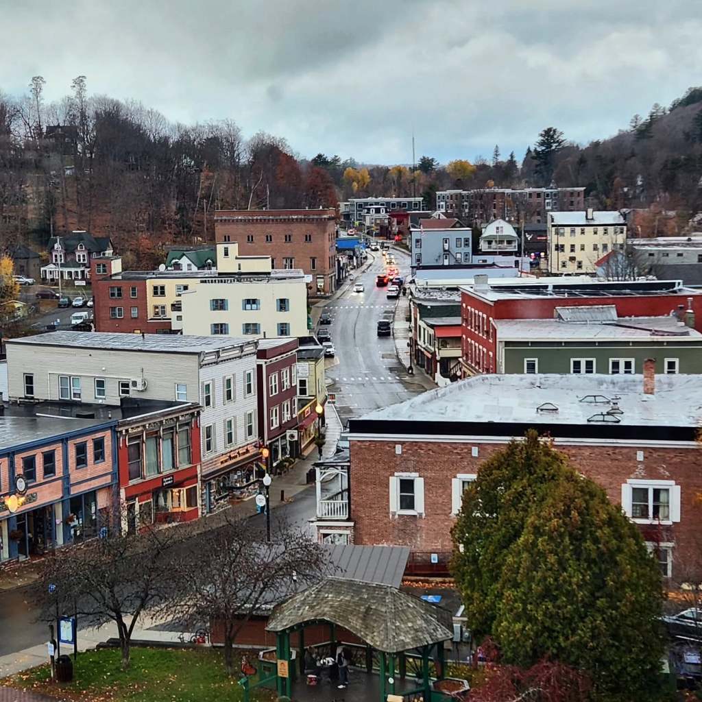 Aerial view of a small town with colorful storefronts and buildings lining a narrow, winding street, under a cloudy sky.