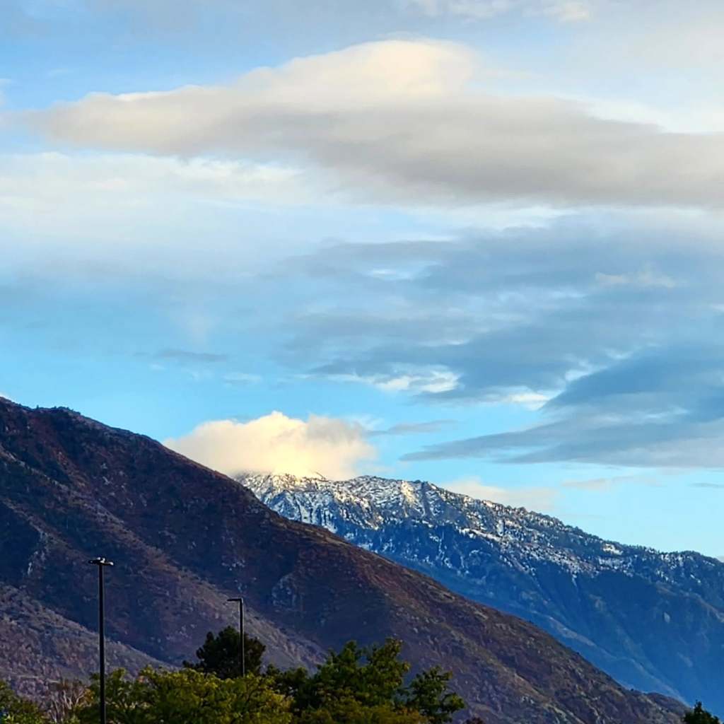 A scenic view of mountains with snow-capped peaks under a partly cloudy sky and greenery in the foreground.