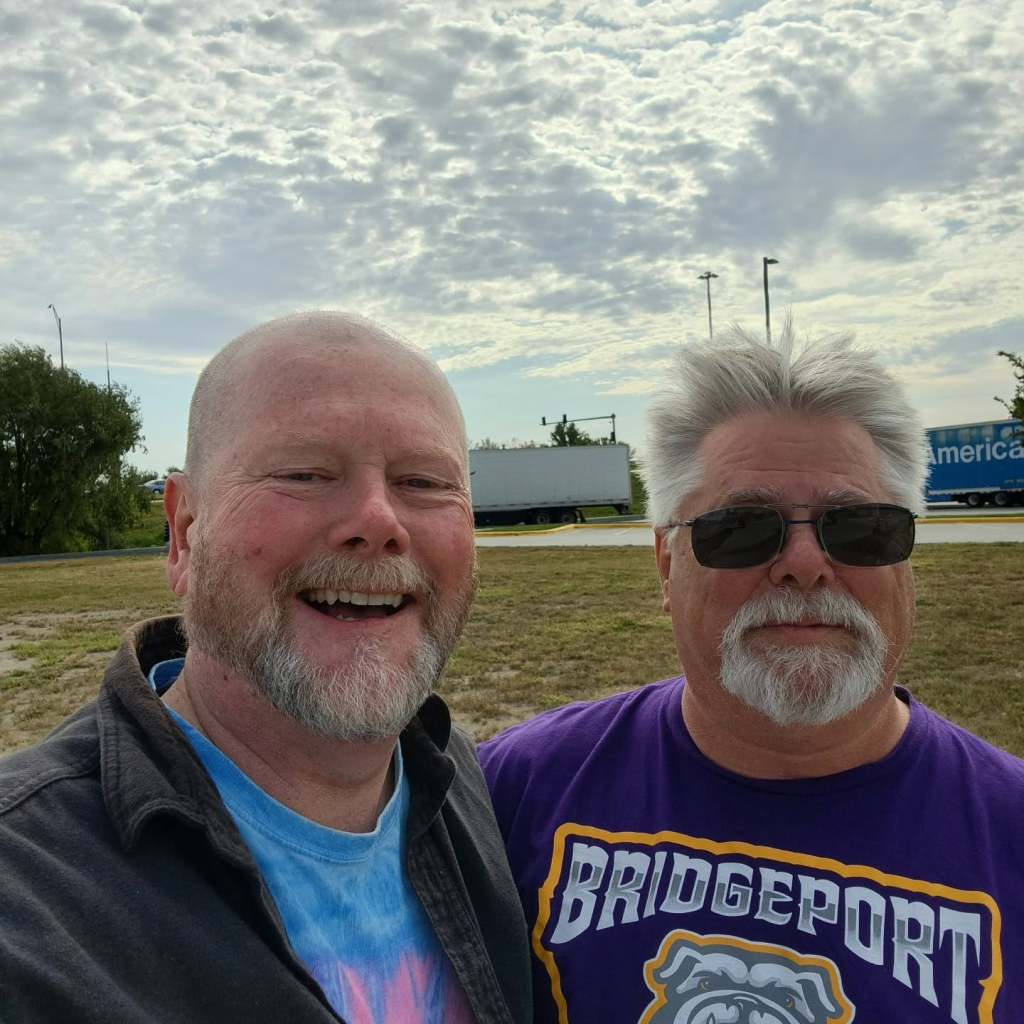 Two men smiling for a selfie outdoors with a cloudy sky in the background. One man has a shaved head and is wearing a colorful shirt, while the other has gray hair and is wearing sunglasses with a purple 'BRIDGEPORT' shirt.